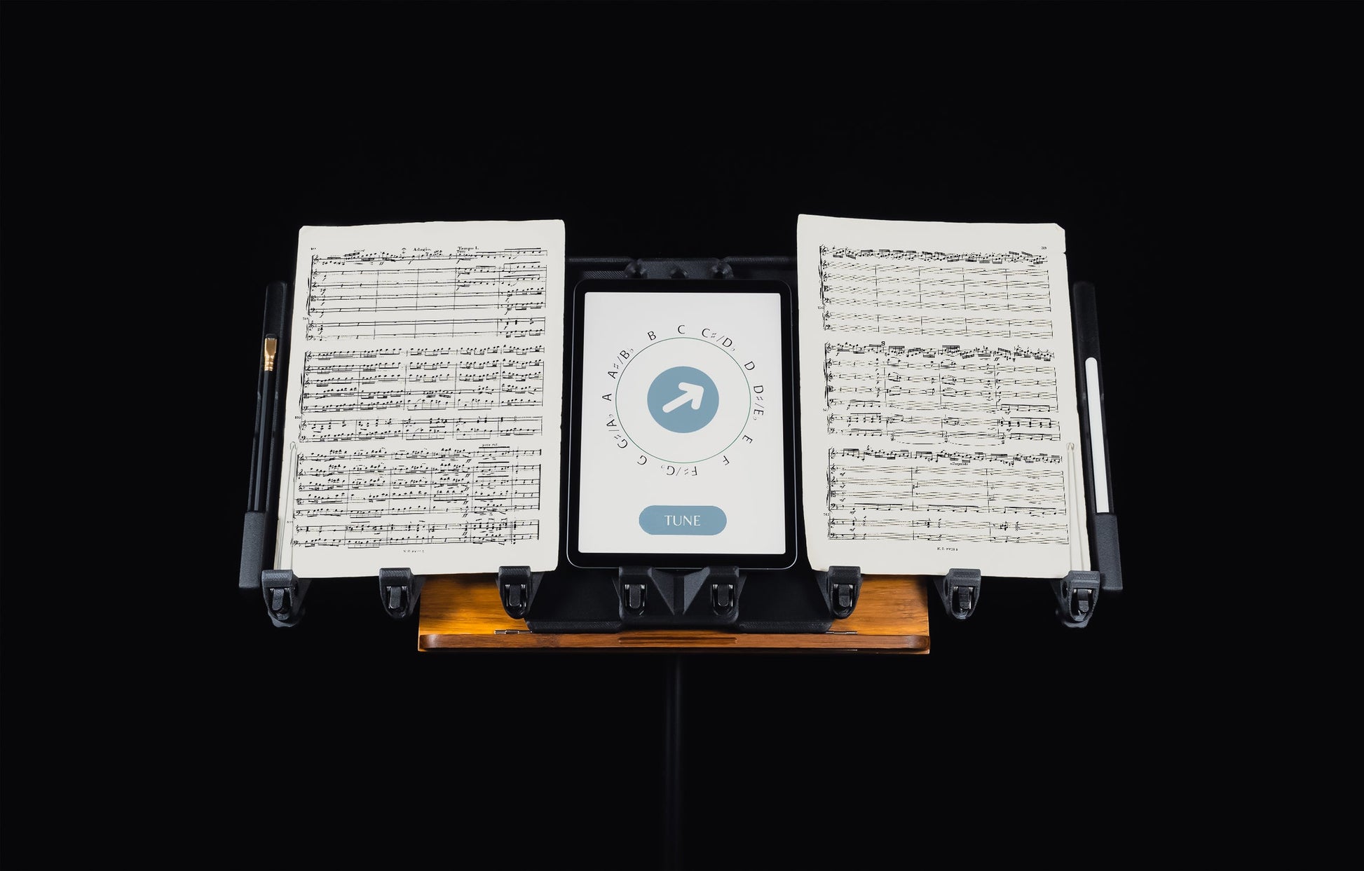 LONGA: Displaying LONGA extending the capabilities of a wooden music stand holding two music pages, a tablet displaying a tunning app, a pencil and a stylus.