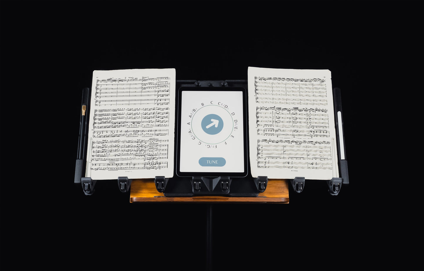 LONGA: Displaying LONGA extending the capabilities of a wooden music stand holding two music pages, a tablet displaying a tunning app, a pencil and a stylus. 